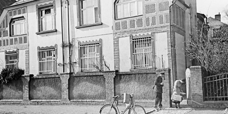 Frontalansicht von der Straße aus auf das Haus 1. Teile des Hauses sind in Fachwerk gehalten. Die unteren Fenster sind alle vergittert. Das Haus hat eine hohe, geziegelte Mauer als Abgrenzung zur Straße. Im rechten Teil ist ein offenes Tor zu sehen. Im Tor steht eine voll bepackte Frau. Neben ihr steht ein Mann mit Schirmmütze und Uniform mit Umhängetasche. Er ist der Frau zugewandt. Auf der Straße vor dem Eingang steht ein Fahrrad an dem Päckchen hängen.