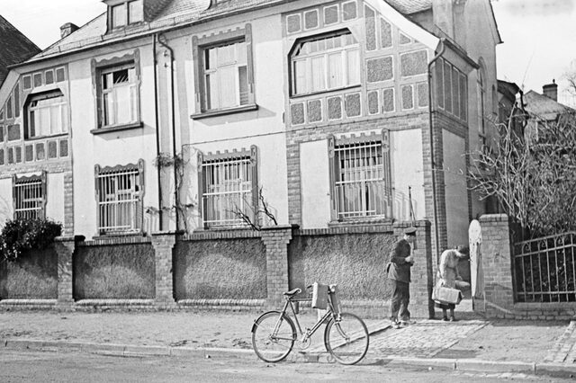 Frontalansicht von der Straße aus auf das Haus 1. Teile des Hauses sind in Fachwerk gehalten. Die unteren Fenster sind alle vergittert. Das Haus hat eine hohe, geziegelte Mauer als Abgrenzung zur Straße. Im rechten Teil ist ein offenes Tor zu sehen. Im Tor steht eine voll bepackte Frau. Neben ihr steht ein Mann mit Schirmmütze und Uniform mit Umhängetasche. Er ist der Frau zugewandt. Auf der Straße vor dem Eingang steht ein Fahrrad an dem Päckchen hängen.