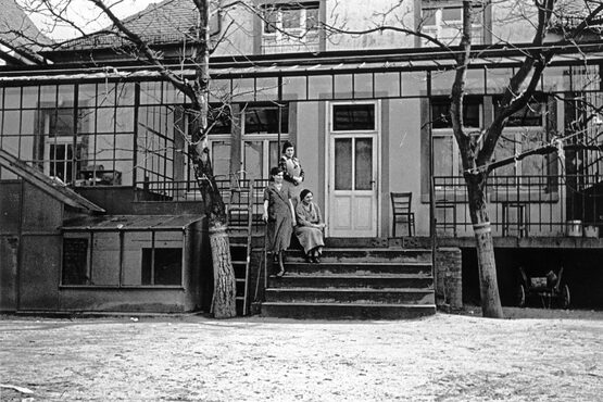 Das Bild zeigt die rückwärtige Seite mit Hinterhof des Haus 1. In der Mitte des Bildes geht eine breite Treppe mit 5 Stufen auf eine überdachte Terrasse. Links und rechts von der Treppe stehen zwei mittelgroße Bäume. Im linken Teil der Terrasse ist ein Wintergarten vorhanden. Zwei Frauen stehen auf unterschiedlichen Ebenen auf der linken Seite der Treppe angelehnt an das Geländer. Eine Frau sitzt daneben auf einer Stufe. Die Blicke der drei Frauen ist in die Ferne gerichtet.