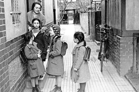 Vier Mädchen stehen mit Mantel und Schulranzen vor einem Hauseingang. Eine Frau steht in der Haustür, hat ihre Hände auf die Schultern eines der Mädchen gelegt und spricht zu den anderen Mädchen.