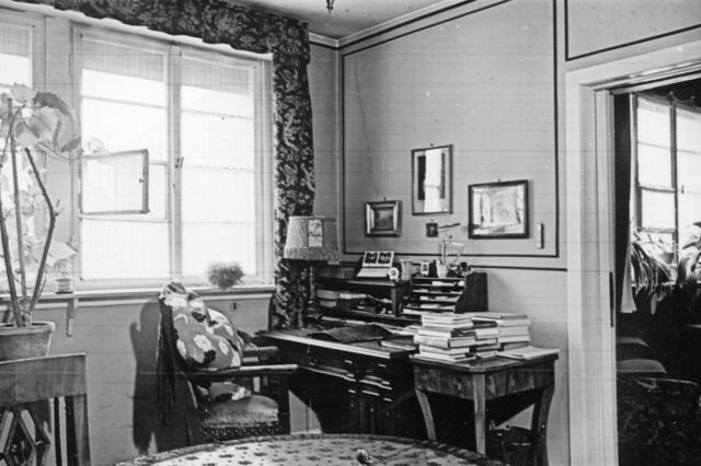 Blick in ein Arbeitszimmer mit einem Sekretär an der Wand. Links ist ein großes Fenster, ein Stück Fenster steht offen. Rechts neben dem Schreibtisch ist ein Durchgang mit einer Schiebetür in ein weiteres Zimmer. Auf dem Sekretär liegen Unterlafen. Links auf dem Sekretär steht eine kleine Stehlampe. Auf einem Beistelltisch rechts neben dem Sekretär sind Bücherstapel. An der Wand hängen Bilder. Links im Bild ist eine Zimmerpflanze zu sehen, die auf einem Tisch steht. Ein Stuhl mit Armlehnen steht schräg vor dem Sekretär.