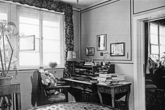 Blick in ein Arbeitszimmer mit einem Sekretär an der Wand. Links ist ein großes Fenster, ein Stück Fenster steht offen. Rechts neben dem Schreibtisch ist ein Durchgang mit einer Schiebetür in ein weiteres Zimmer. Auf dem Sekretär liegen Unterlafen. Links auf dem Sekretär steht eine kleine Stehlampe. Auf einem Beistelltisch rechts neben dem Sekretär sind Bücherstapel. An der Wand hängen Bilder. Links im Bild ist eine Zimmerpflanze zu sehen, die auf einem Tisch steht. Ein Stuhl mit Armlehnen steht schräg vor dem Sekretär.
