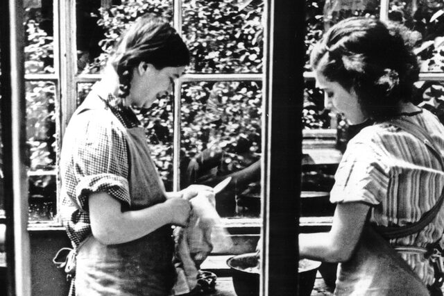 Zwei junge Frauen sind dabei zu sehen, wie sie den Abwasch machen. Die eine hält ein Messer in der Hand, welches sie mit einem Handtuch abtrocknet. Die andere wäscht etwas in einer Schüssel. Die jungen Frauen haben Schürzen an und stehen vor einer großen Fensterfront.