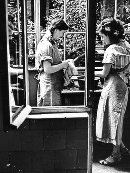 Zwei junge Frauen sind durch ein geöffnetes Fenster und einer Terrassentür dabei zu sehen, wie sie den Abwasch machen. Die eine hält ein Messer in der Hand, welches sie mit einem Handtuch abtrocknet. Die andere wäscht etwas in einer Schüssel. Die jungen Frauen haben Schürzen an und stehen im Wintergarten.