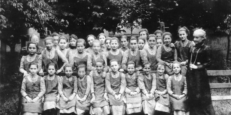 Gruppenfoto von jungen Mädchen, die in drei Reihen hintereinander stehen. Die vorderste Reihe sitzt auf einer Bank. Alle Mädchen tragen die gleiche Kleidung: Ein hochgeschlossenes, helles Unterkleid mit einem farblich abgesetzten, ärmellosen Überkleid. Bertha Pappenheim steht rechts neben der Gruppe sie träft ein dunkles, hochgeschlossenes Kleid mit einer langen Kette.