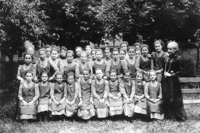 Gruppenfoto von jungen Mädchen, die in drei Reihen hintereinander stehen. Die vorderste Reihe sitzt auf einer Bank. Alle Mädchen tragen die gleiche Kleidung: Ein hochgeschlossenes, helles Unterkleid mit einem farblich abgesetzten, ärmellosen Überkleid. Bertha Pappenheim steht rechts neben der Gruppe sie träft ein dunkles, hochgeschlossenes Kleid mit einer langen Kette.