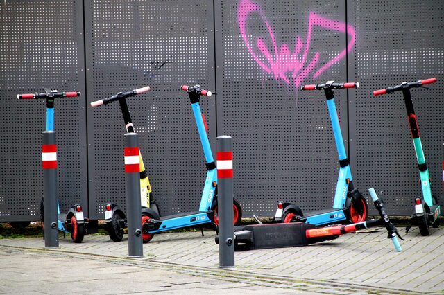 mehrere blaue E-Scooter stehen und liegen auf einem Platz vor einem grauen Metallzaun