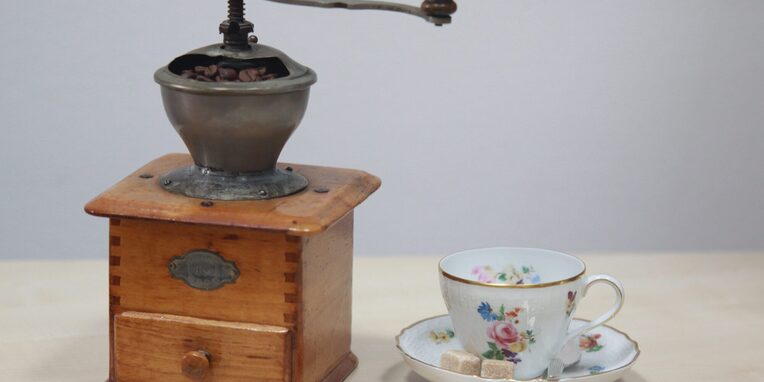 Eine alte braune Kaffeemühle zum drehen mit einer alten weiß geblümten Kaffeetasse und zwei Stückchen Würfelzucker auf dem Unterteller