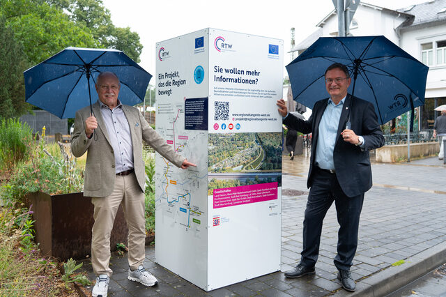 Infostele mit Horst Amann links und Bürgermeister Dirk Gene Hagelstein rechts. Beide halten einen Regenschirm.