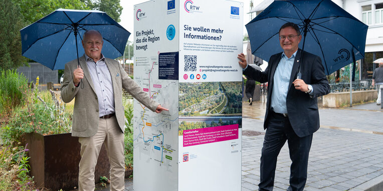 Infostele mit Horst Amann links und Bürgermeister Dirk Gene Hagelstein rechts. Beide halten einen Regenschirm.