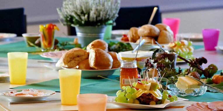 Ein gedeckter, farbenfroher Frühstückstisch mit Brötchen, Butter, Marmelade und Obstvariationen