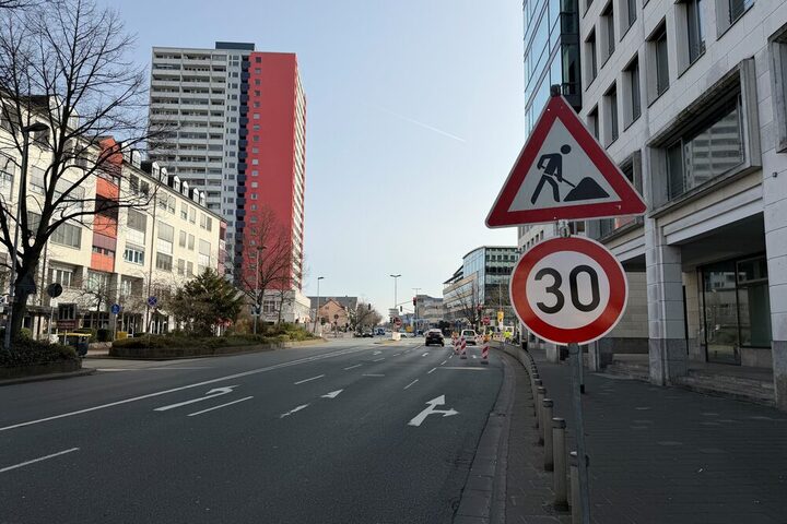 Blick auf die Kreuzung. Rechts: ein rotes Dreieck-Warnschild mit dem Symbol eines Arbeiters (Baustelle) und ein rundes weißes Schild mit rotem Rand und der Zahl 30 (Geschwindigkeitsbegrenzung 30 km/h).