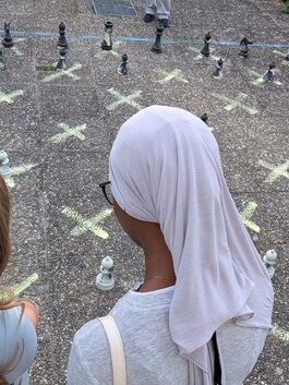 Straßenschach auf die Erde gemalt, im Vordergrund zwei Mädchen von hinten, eines trägt ein Kopftuch